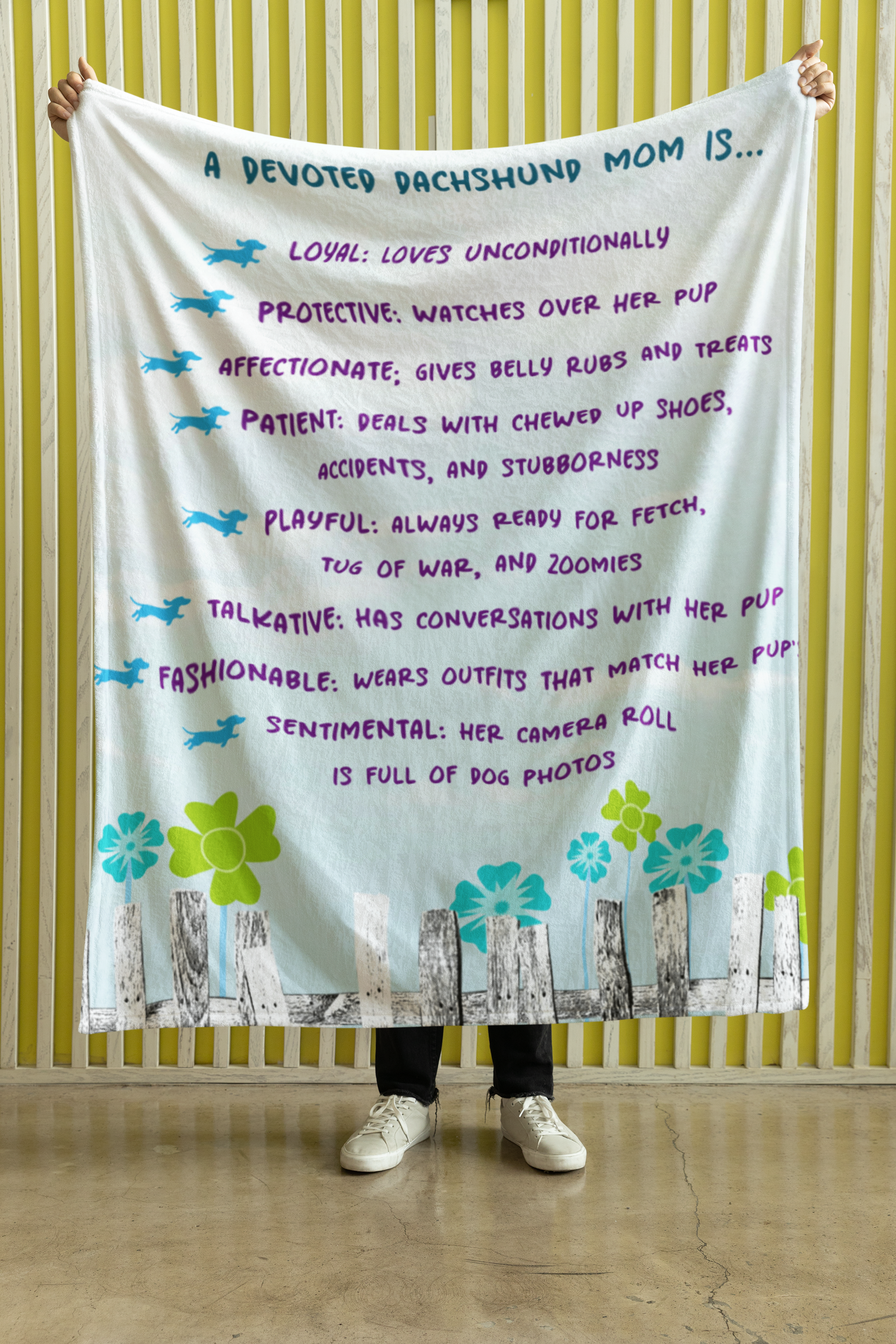 A blanket with text that lists qualities of a devoted dachshund mom, decorated with illustrations of dachshunds, flowers, and a fence.